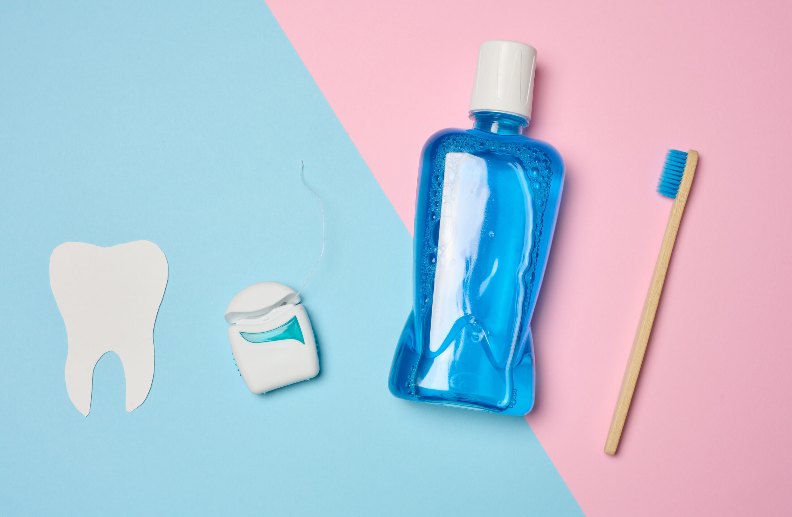 Blue mouthwash bottle, bamboo toothbrush, and dental floss on a pastel pink and blue background.
