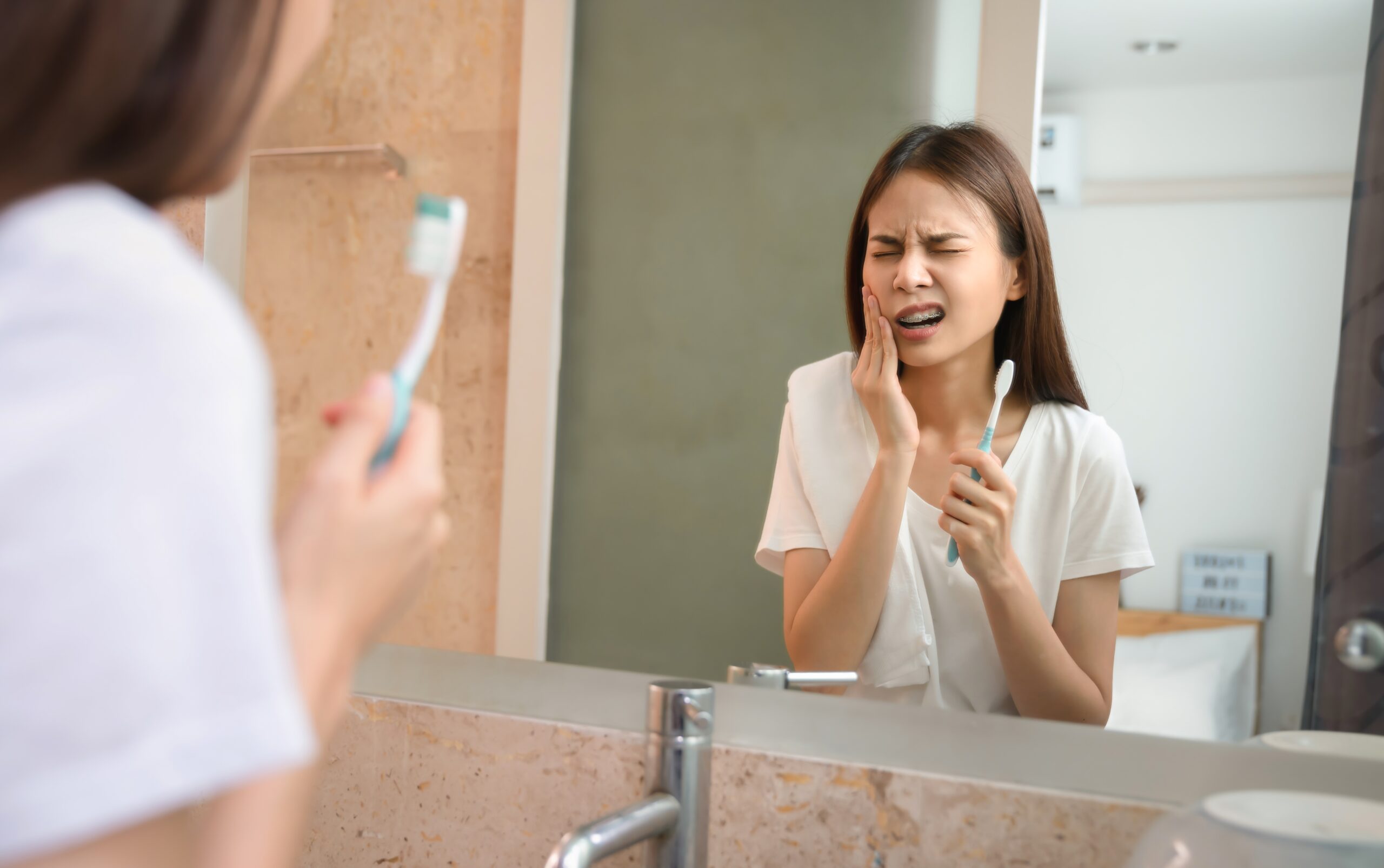 Young woman experiencing tooth sensitivity pain while brushing her teeth in front of a bathroom mirror.