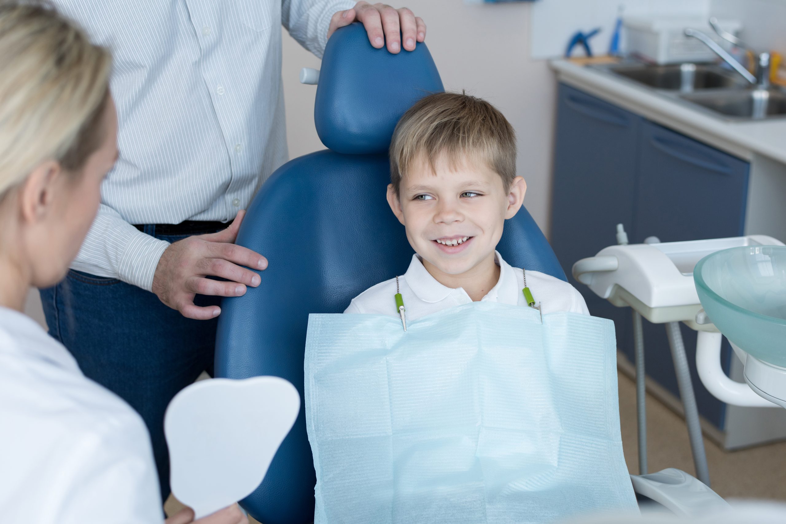 Smiling young boy sitting in a dental chair during his first dental visit, accompanied by a parent and a dentist.