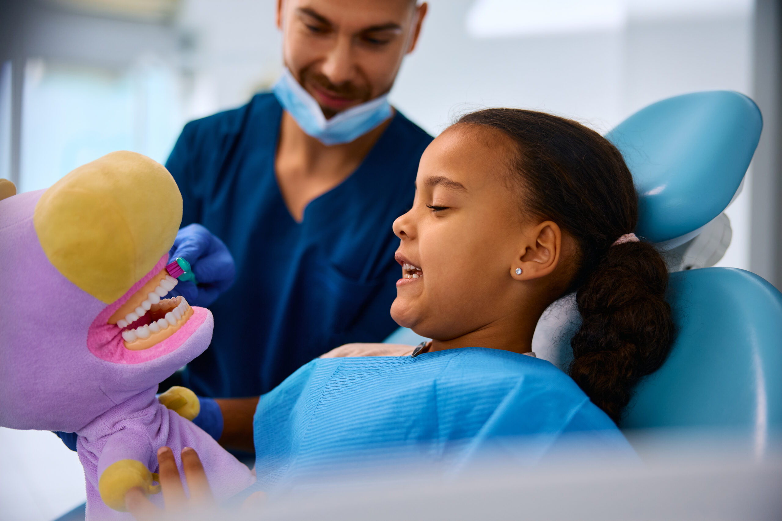A pediatric dentist using a plush toy to teach a young girl how to brush her teeth during a dental visit.