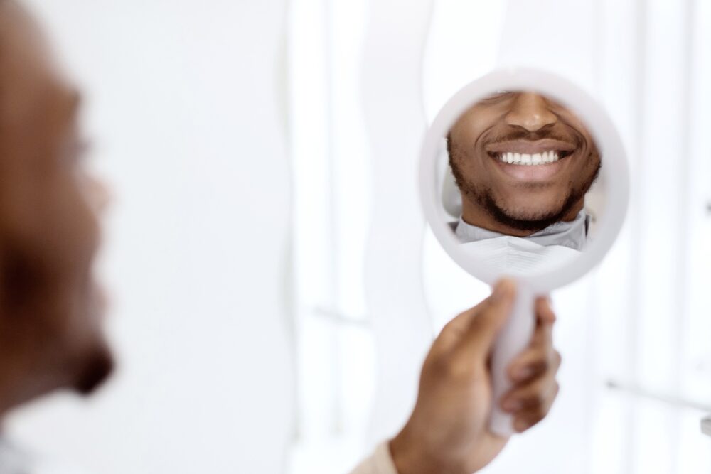 Man smiling into a handheld mirror, showing bright white teeth after whitening treatment